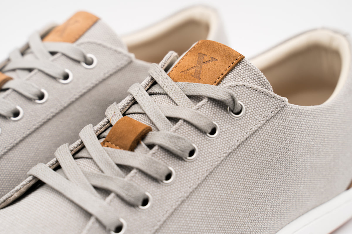 Close-up of a light gray canvas sneaker with matching gray laces, brown leather tongue and lace-loop accents, and a white sole edge on a clean white background.