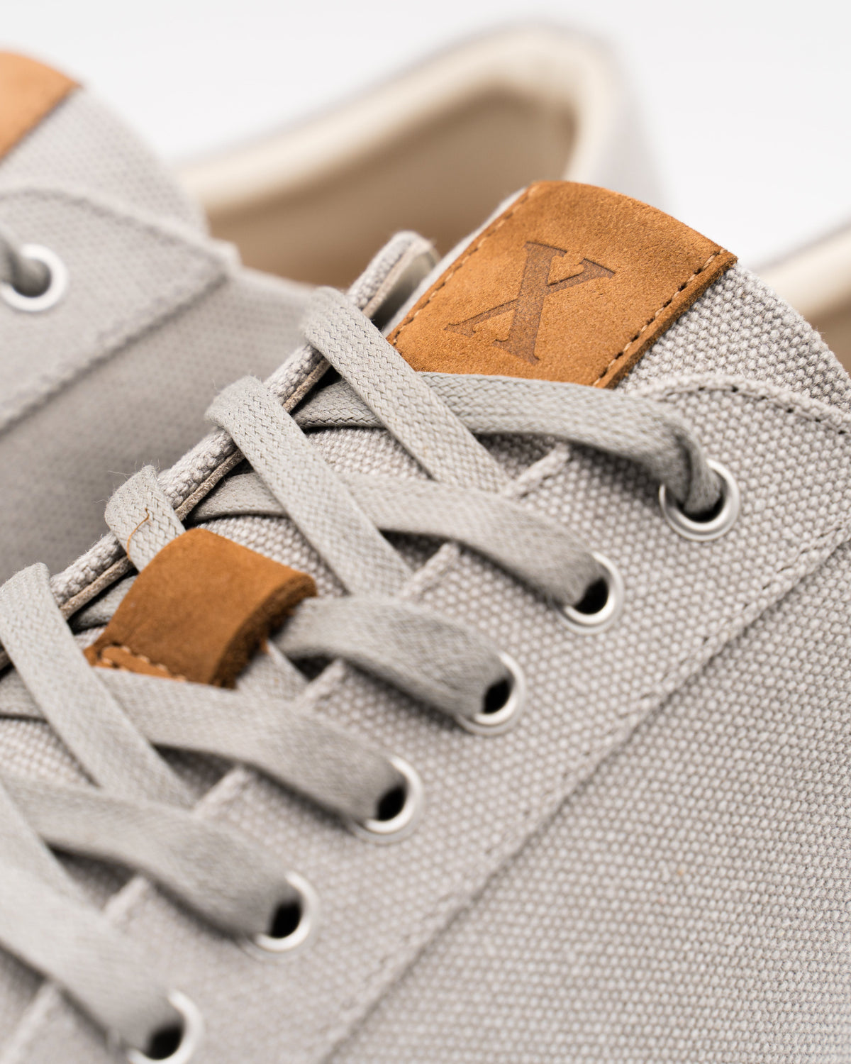 Close-up of a light gray canvas sneaker with matching gray laces, brown leather tongue and lace-loop accents, and a white sole edge on a clean white background.
